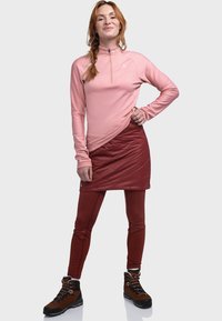 Hellrosa Langarmshirt mit Reißverschlusskragen, rote isolierte Rock, rote Leggings und braune Wanderschuhe mit schwarzen Sohlen und silbernen Akzenten.