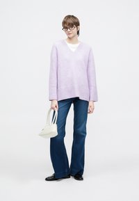 Odvečna vijolična pletena jopica z V-izrezom in rebrastimi manšetami, v kombinaciji s temno modrimi razširjenimi kavbojkami. Drži majhno belo torbico.