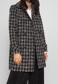 Svart och vit rutig kappa med stora knappar, skräddarsydd passform, bred krage och strukturerad tygdesign, kombinerad med en mörk plisserad nedre del.