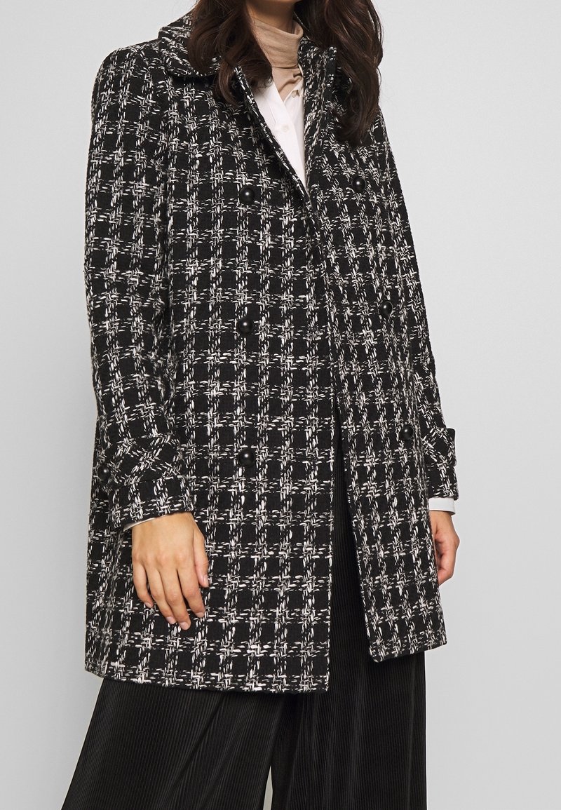 Svart och vit rutig kappa med stora knappar, skräddarsydd passform, bred krage och strukturerad tygdesign, kombinerad med en mörk plisserad nedre del.