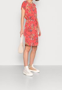 Koralfarvet kjole med blomsterprint, korte ærmer, snørret talje og sideslids. Kombineret med beige sneakers og en tekstureret cremefarvet håndtaske.