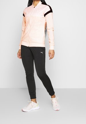 Mujer vestida con una chaqueta rosa claro con cremallera y detalles negros en los hombros, pantalones deportivos negros Puma y zapatos deportivos blancos, de pie frente a un fondo gris.