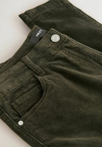 Pantaloni din velur côtelat de culoare verde măslin, cu croială clasică, buzunare frontale și închidere cu buton metalic, având eticheta "NEXT" pe talie.