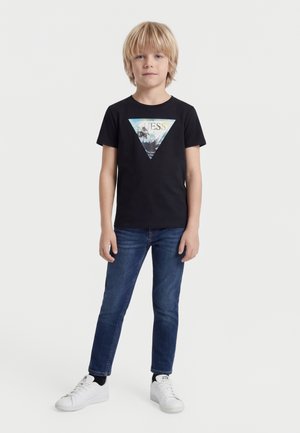 Ένα ξανθό αγόρι στέκεται μπροστά, φοράει μαύρο μπλουζάκι με γραφικό τρίγωνο και μπλε τζιν, λευκά αθλητικά παπούτσια, πάνω σε απλό λευκό φόντο.