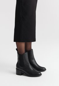 Czarne skórzane botki za kostkę o teksturowanym wykończeniu, z elastycznymi bocznymi panelami oraz grubym obcasem, zestawione z czarną spódnicą.