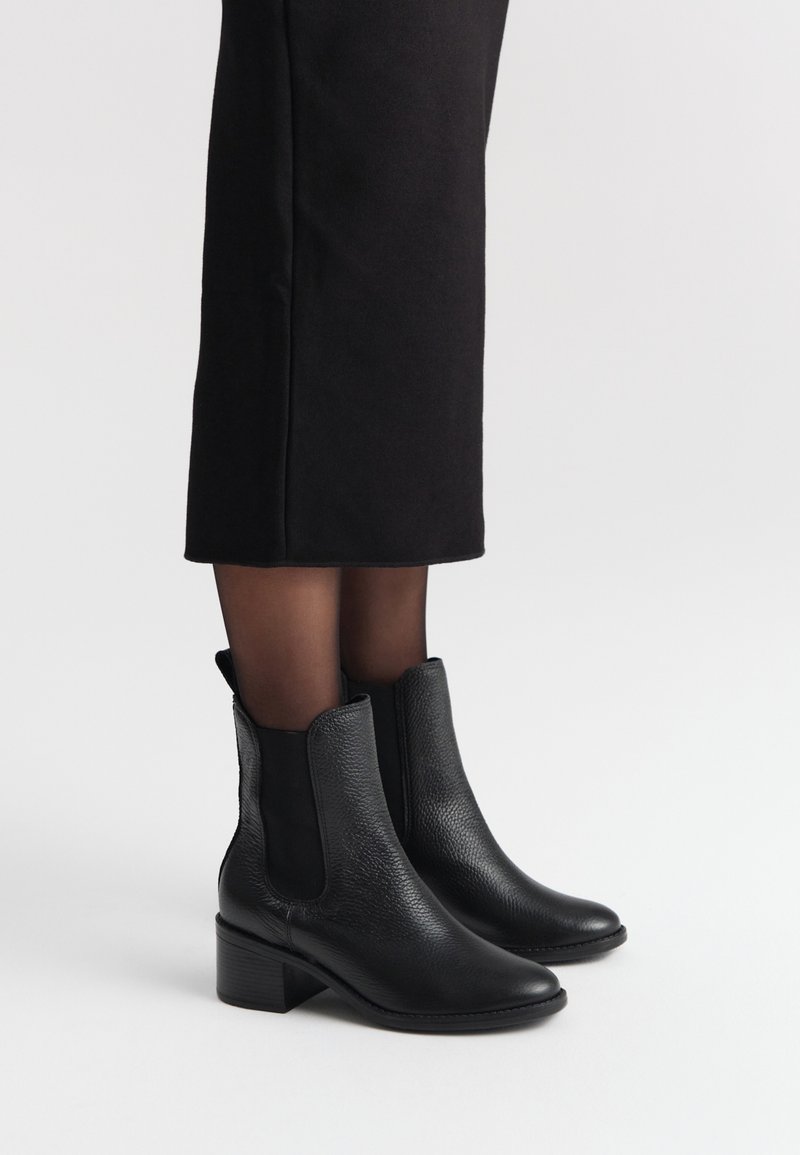 Czarne skórzane botki za kostkę o teksturowanym wykończeniu, z elastycznymi bocznymi panelami oraz grubym obcasem, zestawione z czarną spódnicą.