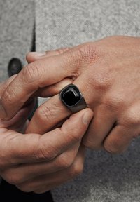 Svart firkantet signetring laget av polert metall, med en blank svart stein innfelt. Hånd plassert med fingrene overlapping.