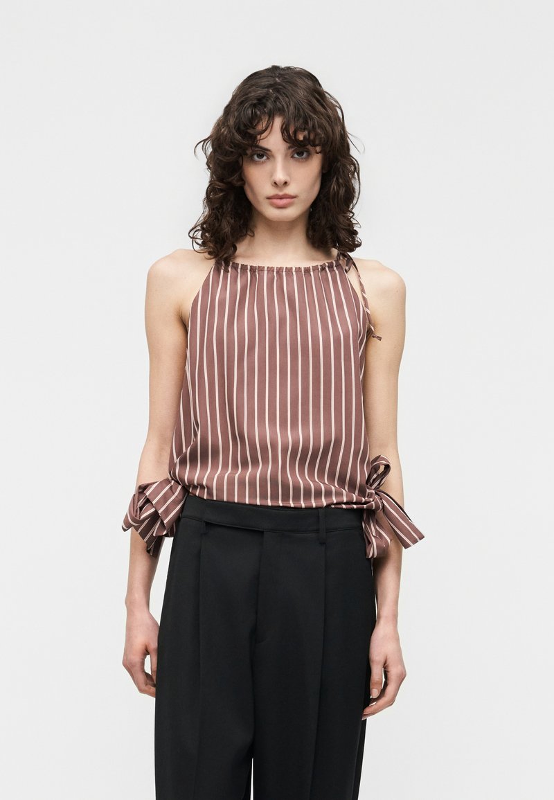 Γυναίκα με σγουρά μαλλιά που φοράει ένα αμάνικο καφέ τοπ με λευκές κάθετες ρίγες και δεμένα φιόγκους στα πλάγια, σε συνδυασμό με μαύρο παντελόνι.