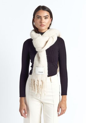 Nainen, jolla on musta ribbineulottu pitkähihaisen paita, valkoiset korkeavyötäröiset housut ja paksu kermaisen värinen huivi, jossa on hapsutetut päät, kietoutuneena kaulan ympärille.