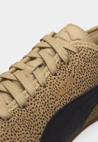 Μπεζ αθλητικό παπούτσι με μαύρο ανάγλυφο σχέδιο λεοπάρδαλης, μπεζ κορδόνια και μαύρη ρίγα λογότυπου Puma στο πλάι.