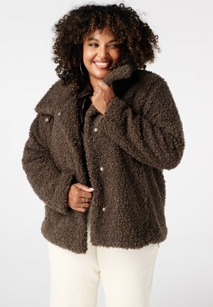 Bruine fluweelachtige jas van een textuur die doet denken aan teddy, met een grote kraag en drukknoopsluiting, gecombineerd met crèmekleurige broeken.