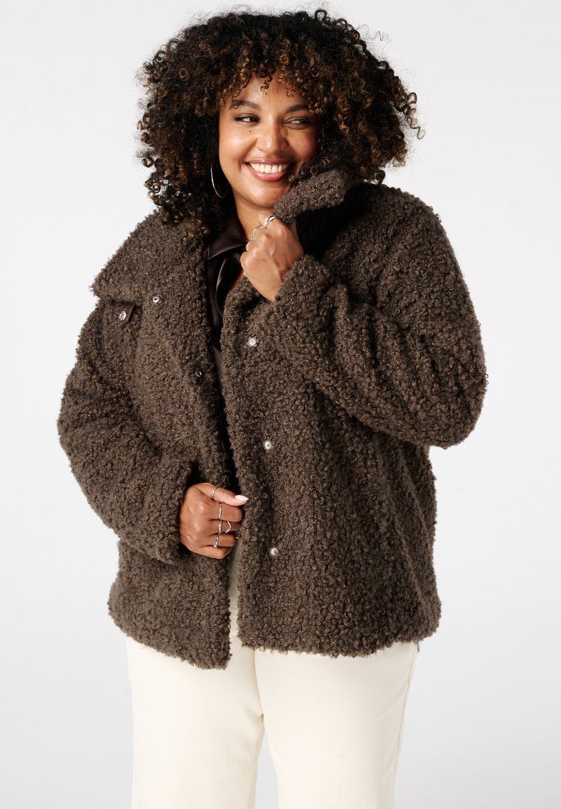 Bruine fluweelachtige jas van een textuur die doet denken aan teddy, met een grote kraag en drukknoopsluiting, gecombineerd met crèmekleurige broeken.