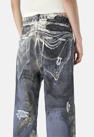 Personne portant un jean taille haute à coupe ample avec des patchs foncés, usés et décolorés sur les poches arrière et les jambes, associé à un haut blanc.