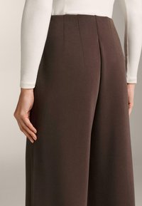 Rjave hlače širokih nog z gladko teksturo in dvema navpičnima šivoma zadaj, v kombinaciji z oprijetim belim topom.