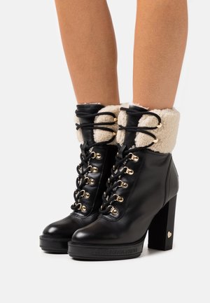 Stivaletti alla caviglia in pelle nera con lacci, tacco spesso e polsini in shearling color crema, indossati su gambe nude.