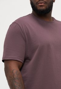 T-Shirt mit kurzen Ärmeln und Rundhalsausschnitt aus einem sanften lila Stoff, der eine glatte Textur und verstärkte Nähte aufweist. Ein Arm ist sichtbar mit einem Tattoo.