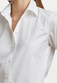 Kvinna i en vit skjorta med knappar och styv krage, som visar överkroppen och halsen mot en enfärgad bakgrund.