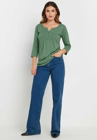 Maglietta verde a maniche lunghe con scollatura henley, caratterizzata da un design plissettato e dettagli con bottoni, abbinata a jeans blu a gamba larga.