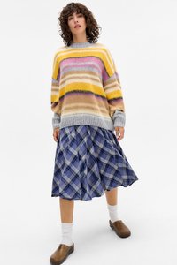 Maglione multicolore a strisce orizzontali gialle, rosa e beige, abbinato a una gonna a quadri blu, indossato con zoccoli marroni e calze bianche.