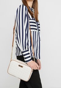 Kobieta w granatowo-białą pionową paskowaną bluzce i czarnych spodniach, nosząca białą torebkę na ramię z brązowym paskiem.