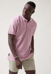 Polo de color rosa claro hecho de algodón, con una tapeta de tres botones y un pequeño logotipo en el pecho; combinado con pantalones cortos beige.