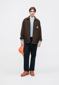 Brunternet blazer over lyseblå sweatshirt, navy bukser, orange kasket og orange sko med teksturerede såler. Model stående mod en hvid baggrund.
