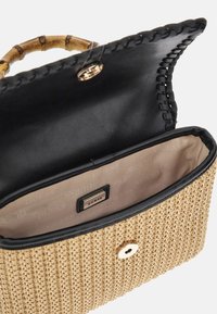 Sac en paille tissé avec un rabat en cuir noir et des ferrures dorées, doté d'une poignée en bambou et d'une doublure beige avec une étiquette de marque à l'intérieur.