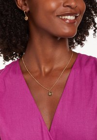 Goldene Halskette mit einem Sonnenanhänger und einer dünnen Kette, kombiniert mit passenden goldenen Creolen, getragen mit einem tiefen V-Ausschnitt in Pink.