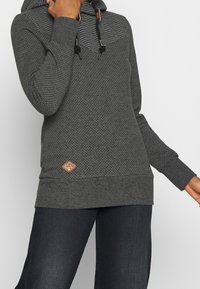 Persoon draagt een donkergrijze hoodie met kleine witte stippen, geribbelde manchetten en een leren merklabel bij de zoom, gecombineerd met een donkere broek.