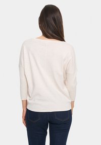 Maglione beige chiaro con scollatura ampia e maniche a tre quarti, con un dettaglio di cucitura orizzontale sul retro. Indossato con jeans scuri.