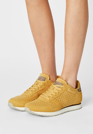 Pieds portant des baskets en mesh et daim jaune moutarde avec des semelles gris clair sur un fond clair uni.