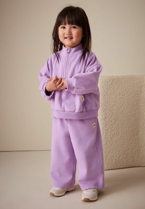 Šviesiai violetinė megzta striukė su užtrauktuku ir derančios kelnės, su ribomis rankogaliuose ir apačioje, su prekės ženklu pažymėtomis etikėtėmis šonuose, dėvint smėlio spalvos sportinius batelius.