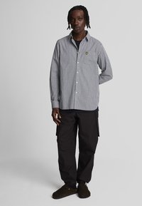 Camicia a quadri in bianco e nero, abbinata a pantaloni cargo scuri e scarpe marroni. Presenta un piccolo logo giallo sul petto.