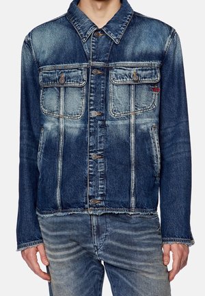 Mann trägt eine ausgewaschene blaue Jeansjacke mit geknöpften Brusttaschen und ausgefransten Kanten, kombiniert mit ausgewaschenen blauen Jeans.