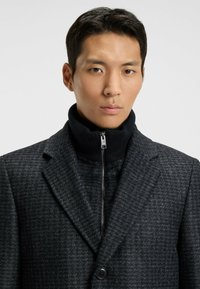 Cappotto grigio a motivi con colletto alto nero con zip, caratterizzato da un tessuto testurizzato e un singolo bottone frontale. Progettato con una vestibilità su misura.