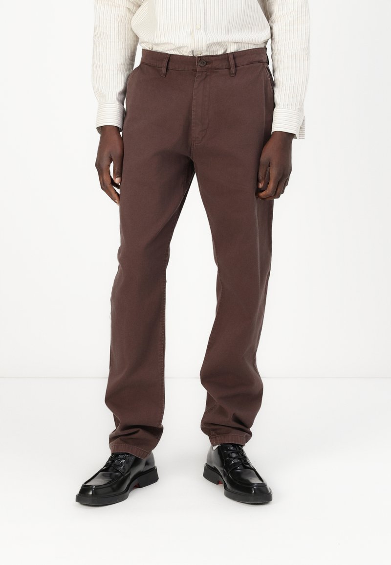 Pantalon en coton brun avec une coupe droite, présentant des poches avant et une fermeture à bouton, associés à des chaussures en cuir noires.