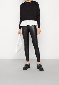 Suéter corto negro con un dobladillo blanco con volantes, combinado con leggins negros brillantes y zapatos negros gruesos, sosteniendo un bolso blanco.