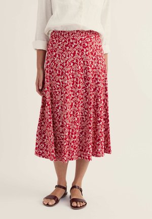 Kobieta ubrana w czerwoną midi spódnicę w biały kwiatowy wzór, białą bluzkę z podwiniętymi rękawami oraz brązowe sandały z paskiem wokół kostki, stojąca na jasnym tle.