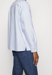 Persona in camicia a maniche lunghe azzurro chiaro e jeans blu scuro, in piedi di lato con la mano rilassata accanto al corpo contro uno sfondo neutro.