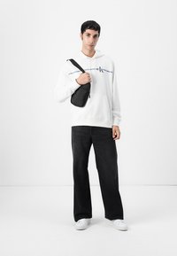 Zwarte schoudertas, witte hoodie met logo aan de voorkant, en donkere wijde jeans. Model draagt witte sneakers en staat tegen een effen achtergrond.