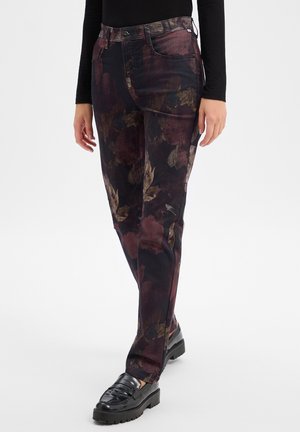 Frau trägt dunkel gemusterte Blumenhose, schwarzen Langarmtop und schwarze Loafers, steht vor einem schlichten Hintergrund.