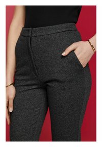 Detailní záběr osoby v tmavě šedých texturovaných kalhotách, se rukou v kapse, černém topu a zlatými náramky na pozadí červené barvy.