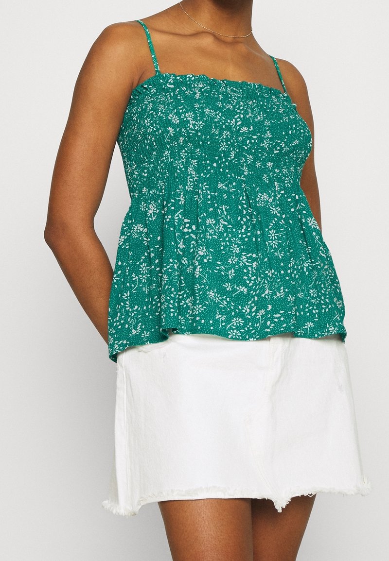 Haut sans manches à motif floral vert avec taille froncée, en tissu léger, associé à une jupe en denim blanc à ourlet brut.