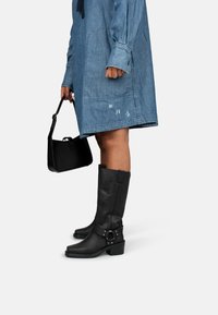 Botas negras hasta la rodilla con un ligero tacón, de materiales de cuero, que presentan un detalle de hebilla lateral. Llevadas con un vestido de mezclilla y sosteniendo un bolso negro.