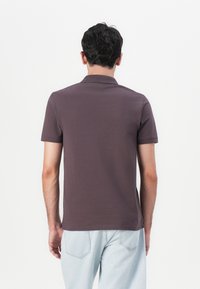 Kurzärmeliges Polohemd aus dunkelbraunem Baumwollstoff. Verfügt über einen flachen Kragen und gerippte Ärmel. Das Modell kombiniert es mit hellblauen Jeans.