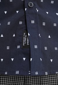 Camicia blu navy con motivi geometrici bianchi, chiusura a bottoni e una piccola etichetta con la scritta "Unified by the Streets" vicino all'orlo.