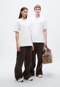 Δύο μοντέλα φορούν λευκά βαμβακερά t-shirt και καφέ παντελόνια με φαρδιά μπατζάκια. Το ένα κρατά μια μπεζ δερμάτινη τσάντα με καφέ λεπτομέρειες. Απλός, καθαρός σχεδιασμός.