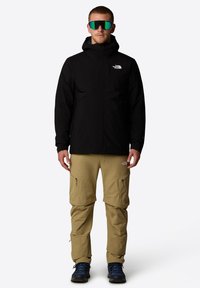 Zwarte waterdichte jas met een rits aan de voorkant en capuchon, gecombineerd met khaki broek met meerdere ritszakken, gedragen met blauwe wandelschoenen.