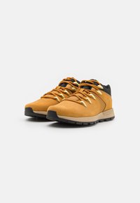 Scarpe da trekking in suede marrone con accenti neri, dotate di occhielli dorati e lacci texture, suola in gomma e colletto imbottito per supporto.