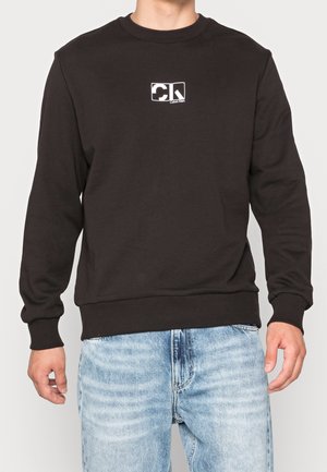 Homme portant un sweat Calvin Klein noir avec un petit logo blanc sur la poitrine et un jean bleu clair, debout devant un fond uni.